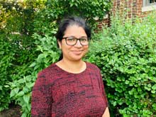 Photograph of staff member Sonam Yadav in front of a green leafy bush. She is smiling, wears glasses and is wearing a red top.