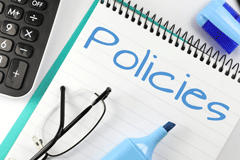 Stock photo of "Policies" written in blue highlighter on a notepad. Notepad is next to a keyboard and has a pair of glasses folded on it.